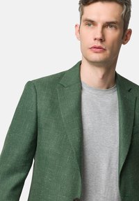 Zielona marynarka w jodełkę z klapkami, jednopunktowym zapięciem, noszona na jasnoszarym T-shircie z okrągłym dekoltem. Gładka tekstura, dopasowany krój.