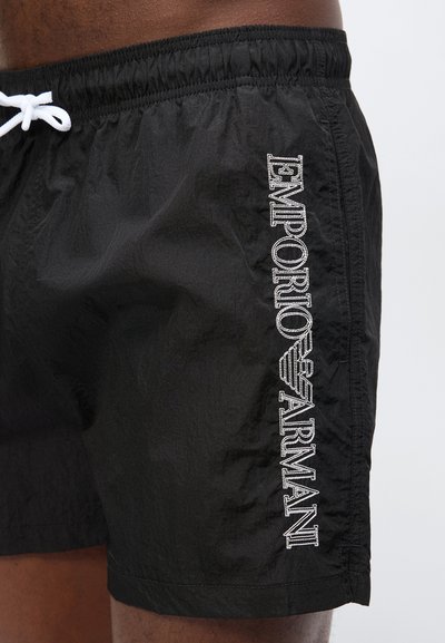 Μαύρα ανδρικά μαγιό βερμούδα με λευκό κορδόνι και λευκό κάθετο λογότυπο "EMPORIO ARMANI" στο πλάι που φοριούνται από ένα άτομο.