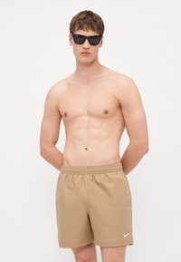 Shorts de bain beiges en tissu lisse avec une ceinture élastique et le logo blanc de Nike. Modèle portant des lunettes de soleil noires, debout avec les mains derrière le dos.