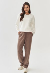 Donna con lunghi capelli castani, che indossa una felpa bianca, pantaloni marroni in maglia con coulisse e scarpe da ginnastica bianche, in piedi con le mani nelle tasche.
