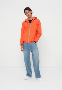 Chaqueta ligera de color naranja brillante con cierre de cremallera, detalles en azul marino, capucha y ajuste relajado, combinada con unos vaqueros de pierna ancha de color azul claro.