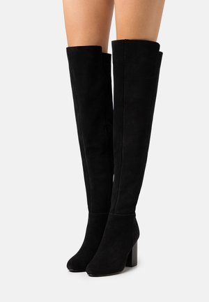 Mujer vistiendo botas de ante negras hasta la rodilla con tacones cuadrados contra un fondo blanco liso.