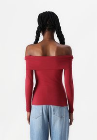 Raudonas off-the-shoulder ilgas rankovės viršus su prigludusia forma, lygiu paviršiumi ir kontrastingomis mėlynomis aukšto liemens džinsomis.