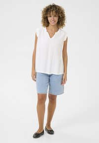 Haut blanc à manches courtes avec plis au niveau du col, associé à un short en denim bleu clair. Des ballerines noires complètent la tenue.