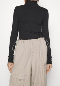 Schwarzes, geripptes Rollkragenoberteil kombiniert mit beigen Cargo-Hosen, die elastische Kordeln, Taschen und geraffte Details an der Taille aufweisen.