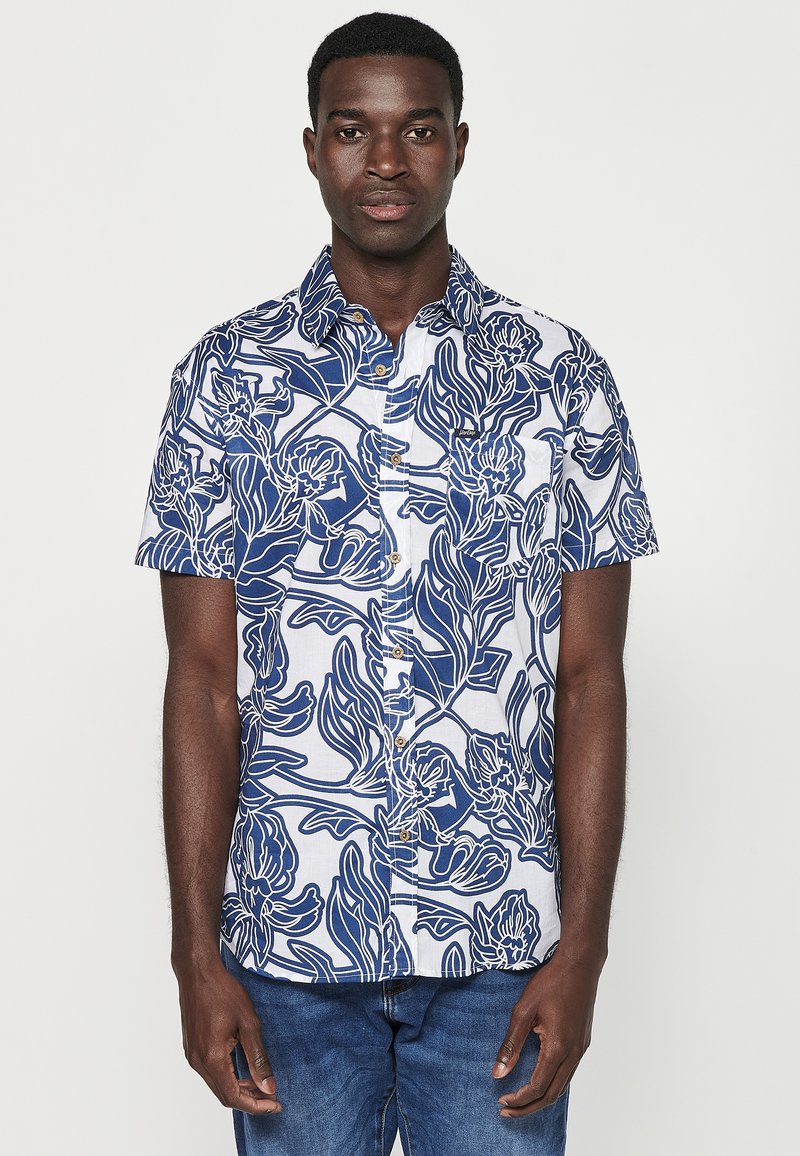 La camicia button-up presenta una base bianca con motivi floreali blu, maniche corte e un colletto. Il tessuto appare leggero e texturizzato.