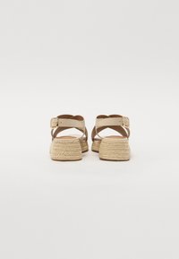 Beige espadrille-sandaalit, joissa on ristitimantti-muotoilu, suljetut takapuolet ja kudotut köysikengät. Kultaiset solkikoristeet säädettävillä remmeillä.