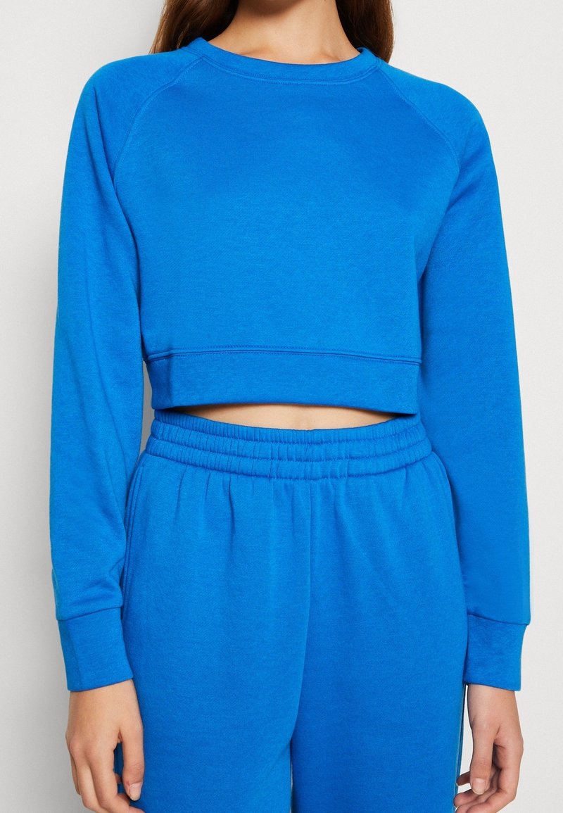 Jeune femme portant un sweat-shirt court à manches longues bleu vif et un pantalon de jogging taille haute assorti avec une ceinture élastique.