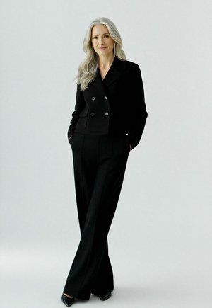 Vrouw met lang grijs haar, gekleed in een zwarte dubbel-geknoopte blazer, brede broek, zwarte puntige schoenen en lange hangende oorbellen, staat ontspannen.