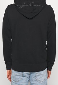 Musta hupullinen collegepaita, jossa on edessä kengurutasku, resorit hihansuissa ja helmassa, ja kontrastivärinen nimikirjaintunnus takana.