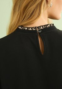 Top nero con dettaglio a chiave sulla schiena, adornato con un volant a stampa leopardata e chiusura con bottone sul collo. Texture liscia.