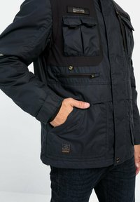 Sort isoleret jakke med flere forlommer, en lynlås lukning og et struktureret stof. Har et logo patch og kontrastpaneler på skuldrene.