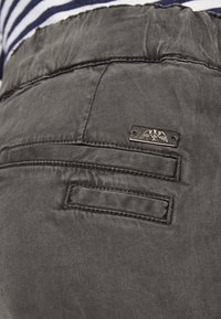 Dunkelgraue Hosen aus einem glatten Material, mit einem elastischen Taillenbund und zwei Gesäßtaschen mit einem metallischen Markenetikett.