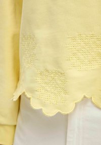 Indumento giallo chiaro con orlo scallop, rifinito da un ricco ricamo a motivo fiocco di neve lungo il bordo inferiore. Tessuto dalla consistenza liscia.