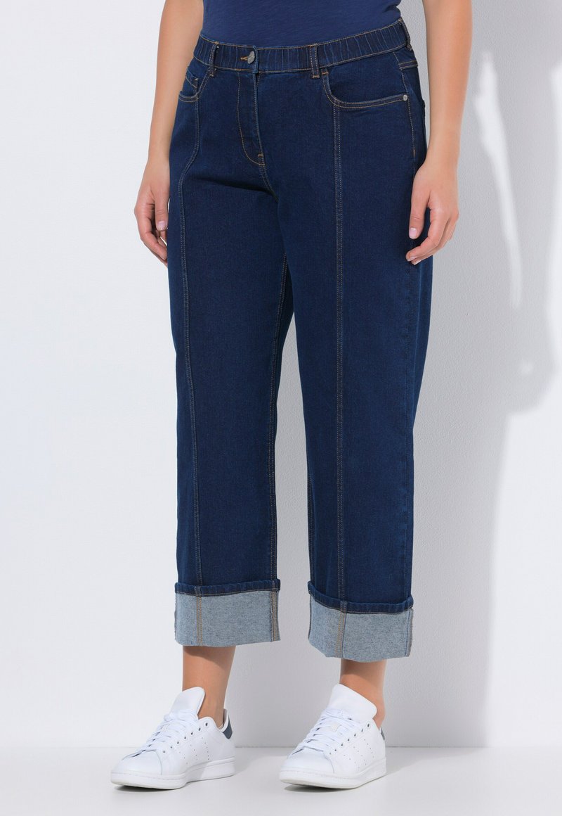 Osoba v tmavě modrých ohrnutých džínách a bílých teniskách stojí před jednobarevným bílým pozadím.