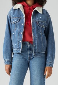 Džínsová bunda s límcem z sherpy, v modrém odstínu, s opotřebovanými detaily a kovovými knoflíky. Nositá přes červenou mikinu.