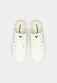 Bílé plátěné tenisky s kulatou špičkou, plochými šňůrkami a logem potisknutým na jazyku. Texturovaná podrážka s minimalistickým designem.