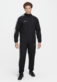 Schwarzer Trainingsanzug aus glattem Material, bestehend aus einer Jacke mit Reißverschluss und grauen Schulterakzenten sowie passenden Hosen, beide geschmückt mit Nike-Logos.