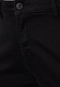 Pantalones de mezclilla negros con una textura suave, que cuentan con un botón metálico visible, bolsillos laterales y costuras de contraste. Vista en primer plano.