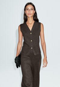 Femme aux longs cheveux noirs portant un gilet sans manches marron foncé boutonné et un pantalon assorti, tenant une pochette noire.