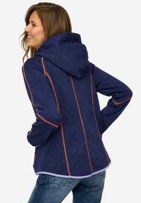 Veste à capuche de couleur marine avec des coutures orange contrastantes, texture douce et ourlet arrondi. Portée par-dessus un jean en denim, montrant un style décontracté.