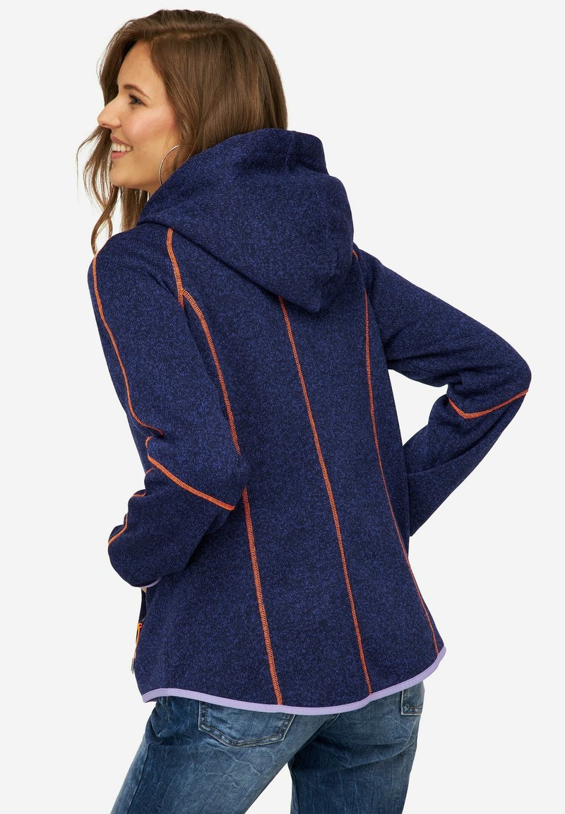 Veste à capuche de couleur marine avec des coutures orange contrastantes, texture douce et ourlet arrondi. Portée par-dessus un jean en denim, montrant un style décontracté.