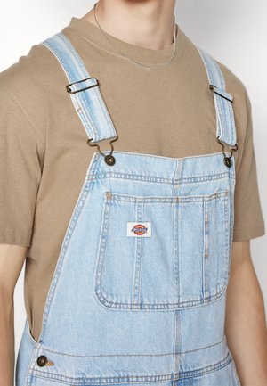 Osoba nosząca jasno niebieski deniomowy ogrodniczki z przednią kieszenią i brązową koszulkę z krótkim rękawem pod spodem, widoczny srebrny łańcuszek.