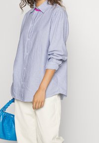 Sininen ja valkoinen raidallinen napillinen paita, rento malli rullattavilla hihoilla, yhdistettynä teksturoituun siniseen käsilaukkuun.