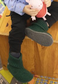 Groene vilten kinderschoenen met een geribbelde boord en een grijze zool, gedragen door een kind dat een knuffel vasthoudt en op een houten oppervlak zit.