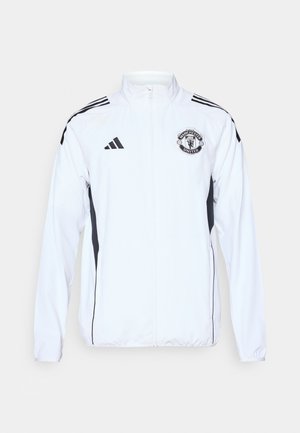 Veste de sport blanche avec des accents noirs, fermeture éclair, et logo de Manchester United. Présente trois bandes noires sur les épaules et des poignets élastiques.