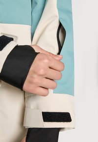 Lightweight outerwear features a color-block design in mint green and cream, with a black elastic cuff and zippered pocket detail.