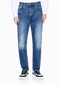 Blaue Jeans aus Denim mit geradem Schnitt, fünf Taschen, dezentem Verblassen und gestickten Akzenten. Getragen mit schwarz-weißen Sneakers.