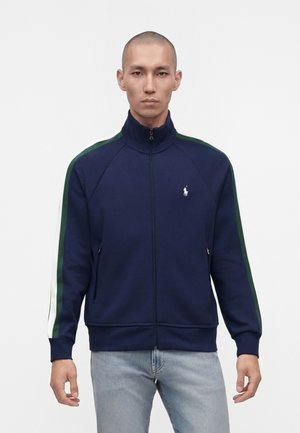 Mann mit rasiertem Kopf, der eine marineblaue Zip-up-Jacke mit grünen und weißen Streifen an den Ärmeln sowie hellblaue Jeans trägt und vor weißem Hintergrund steht.