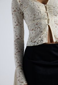 Spitzen-Crop-Top mit Blumenmuster, cremefarben, mit langen Ärmeln, ausgestattet mit drei Perlenknöpfen und einem ausgestellten Ärmel-Design. Schwarze hochtaillierte Röcke.