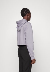 Krótkie lilakowe hoodie z kapturem na sznurku i ściągaczami przy mankietach, pokazane od tyłu, w komplecie z ciemnymi spodniami w kant. Wykonane z miękkiego materiału bawełnianego.
