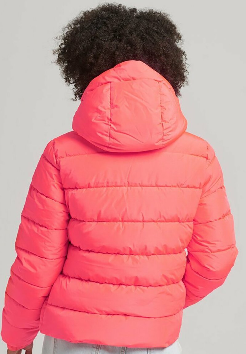 Persona con capelli ricci che indossa una giacca a vento corallo brillante con cappuccio, vista da dietro su uno sfondo grigio uniforme.