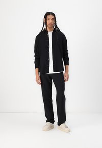 Svart, langermet skjorte med knapper over en hvit t-skjorte, kombinert med svarte bukser og beige sneakers. Minimalistisk design og avslappet passform.