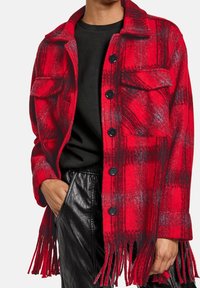 Chaqueta de lana de cuadros rojos y negros con botones negros y dobladillo de flecos, llevada sobre una camisa negra y pantalones de cuero negros.