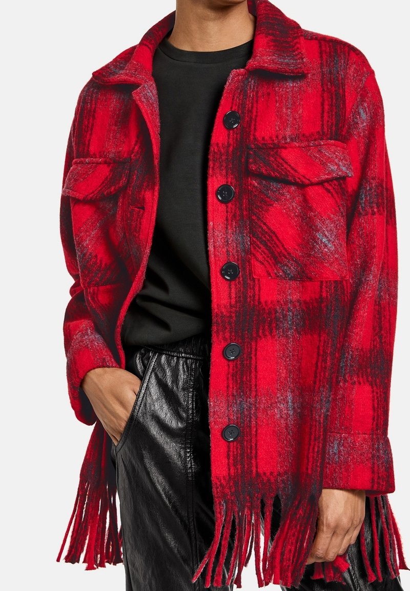 Chaqueta de lana de cuadros rojos y negros con botones negros y dobladillo de flecos, llevada sobre una camisa negra y pantalones de cuero negros.