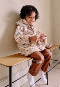 Chaqueta impermeable de color beige claro con estampados de animales coloridos, combinada con pantalones impermeables marrones y calcetines blancos, niño sentado en un banco de madera.