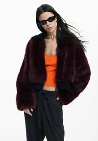 Bordová bomber bunda z umelého kožúšku s tmavomodrým golierom, v kombinácii s oranžovým vrúbkovaným crop topom, čiernymi nohavicami a tmavými slnečnými okuliarmi.