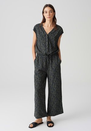 Marineblauer Jumpsuit mit grünem Blumenmuster, kurzen Ärmeln, V-Ausschnitt, elastischem Bund und weit geschnittenen Hosenbeinen. Kombiniert mit schwarzen Rutsch-Sandalen.
