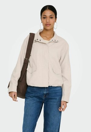 Žena v svetlobé béžovej bunde a modrých džínsoch, nesúca veľkú hnedú kabelku na ramene, s vlasmi stiahnutými dozadu a kruhovými náušnicami.