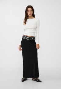 Black maxi skirt paired with a fitted white long-sleeve top, accented with a silver-studded black belt and black pointed-toe shoes.
