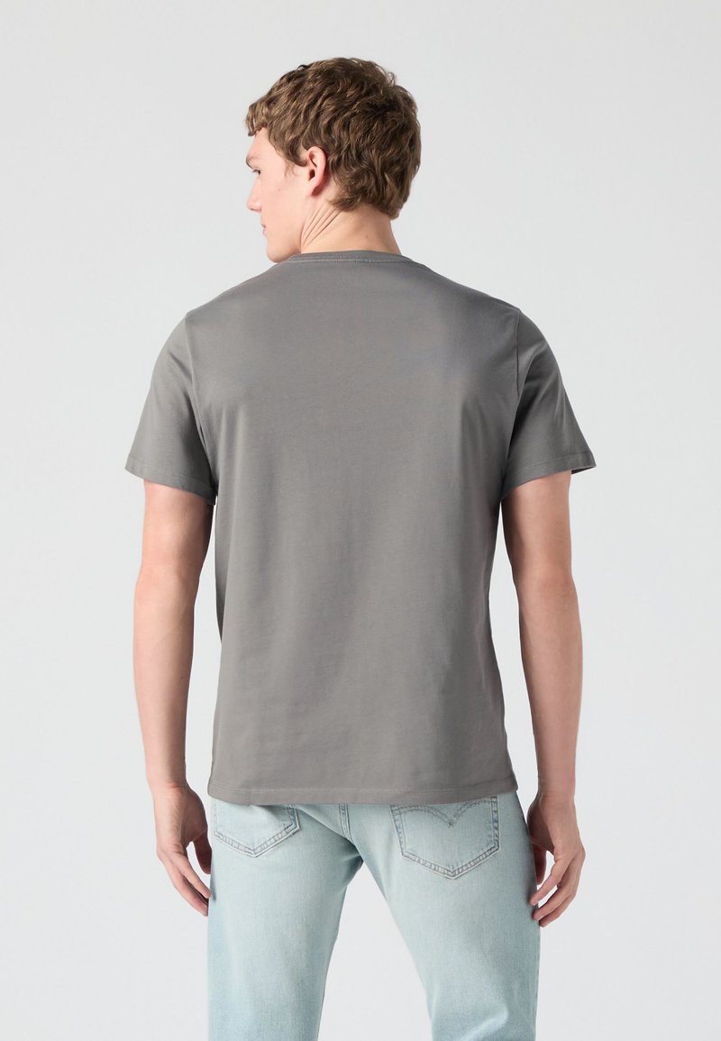 Homme de dos portant un t-shirt gris uni à manches courtes et un jean bleu clair devant un fond uni.