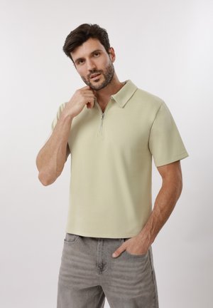 Άνδρας με κοντά σκουρόχρωμα μαλλιά και γένια, φορώντας μπεζ πουκάμισο με κοντά μανίκια και φερμουάρ στη λαιμόκοψη και γκρι τζιν, ποζάροντας με το ένα χέρι κοντά στο πρόσωπο.