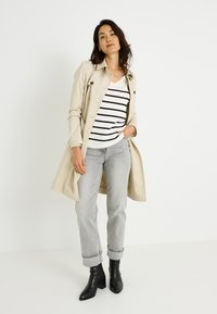 Abrigo trench beige sobre una camisa blanca con rayas negras, combinado con vaqueros gris claro remangados y botines negros.