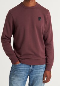 Bordowy sweter z miękkiego materiału, z okrągłym dekoltem, długimi rękawami, ściągaczami na mankietach oraz małą czarną naszywką z logo na klatce piersiowej.