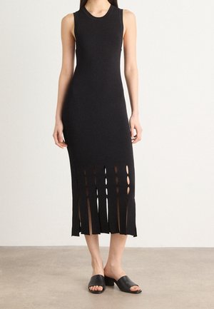 Robe midi noire sans manches avec encolure ronde et fentes verticales à l'ourlet, portée avec des mules à talons noires ouvertes sur un sol neutre.