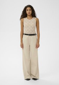 Kobieta z długimi kręconymi włosami, ubrana w bezrękawnik z beżowej koronki, szerokie spodnie z wysokim stanem w beżowym kolorze, czarny pasek i czarne buty, stojąca.